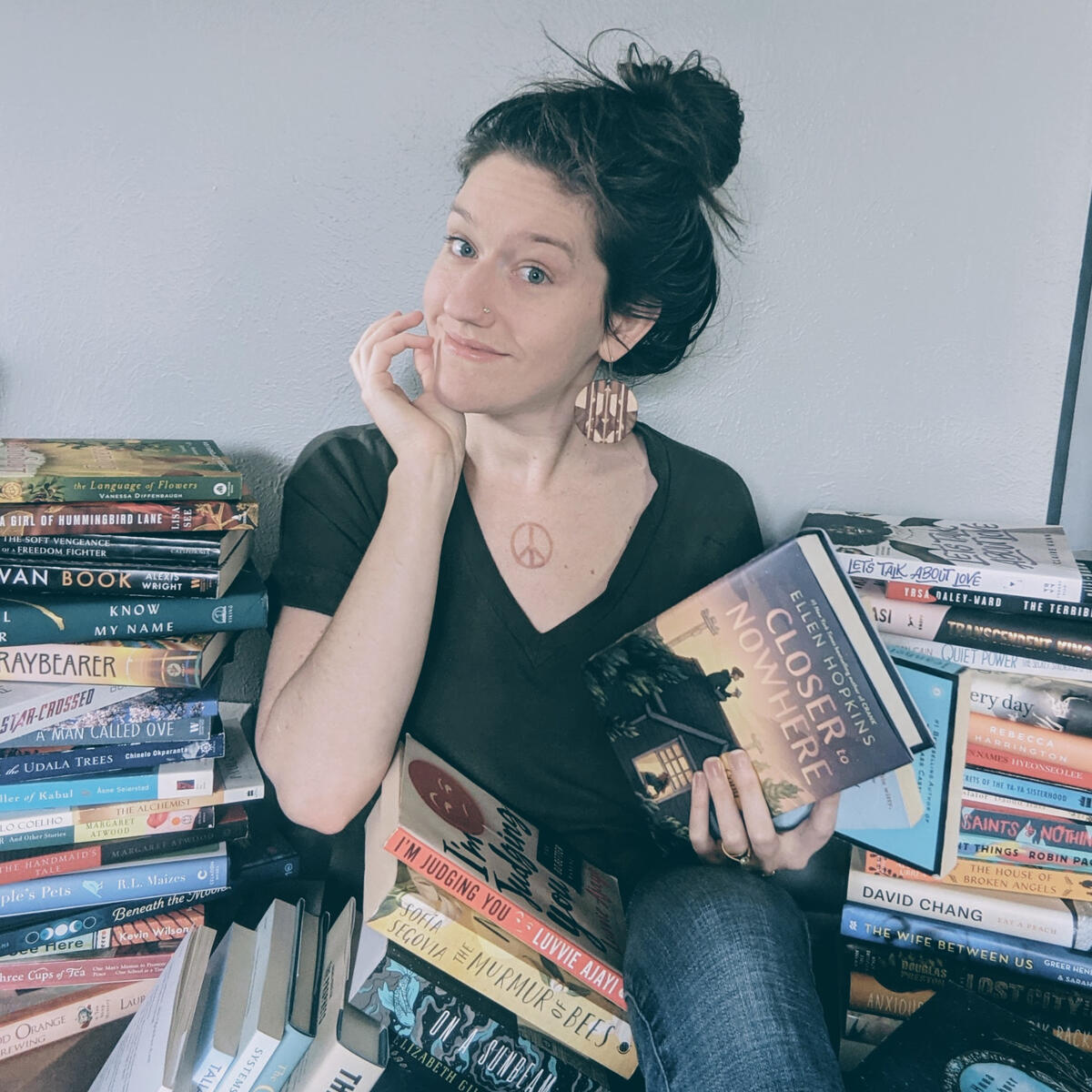 christina sits between 2 large piles of books. one stack of books is falling over on her lap, and she holds 3 books in her hand.
